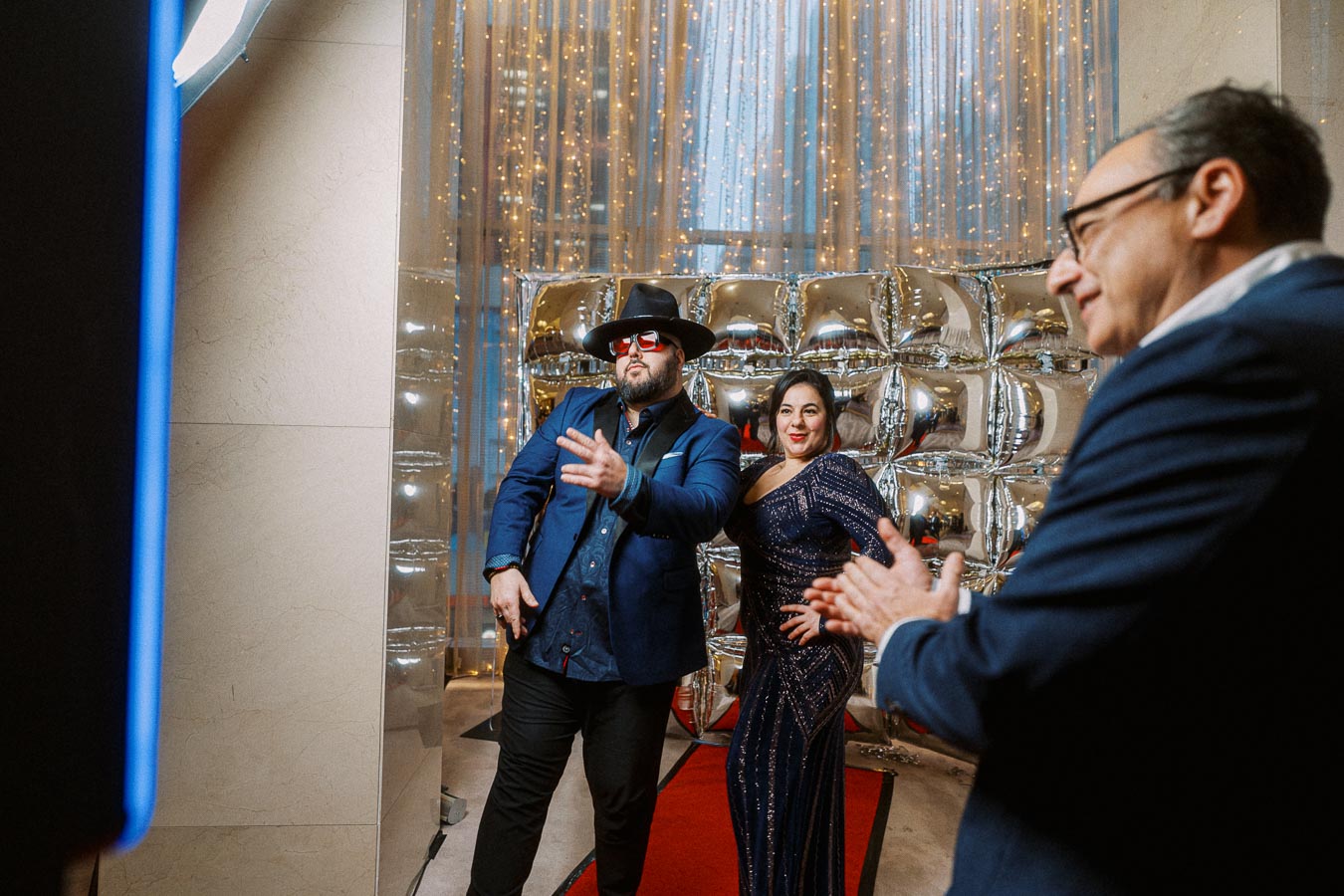 A stylish man and woman pose confidently in front of a glamorous backdrop with silver balloons and twinkling lights, beside a red carpet, as another person looks on in admiration, all dressed in formal attire.
