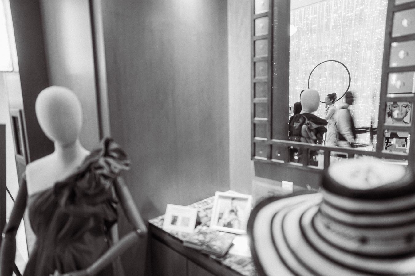Black and white photo of a fashion boutique interior featuring a mannequin dressed in an elegant off-shoulder garment, a stylish hat in the foreground, and a blurred reflection of people in a mirror surrounded by decor and framed pictures.