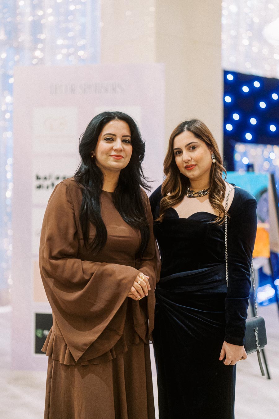 Two women in elegant outfits pose at an indoor event, with blurred decorative lights in the background.