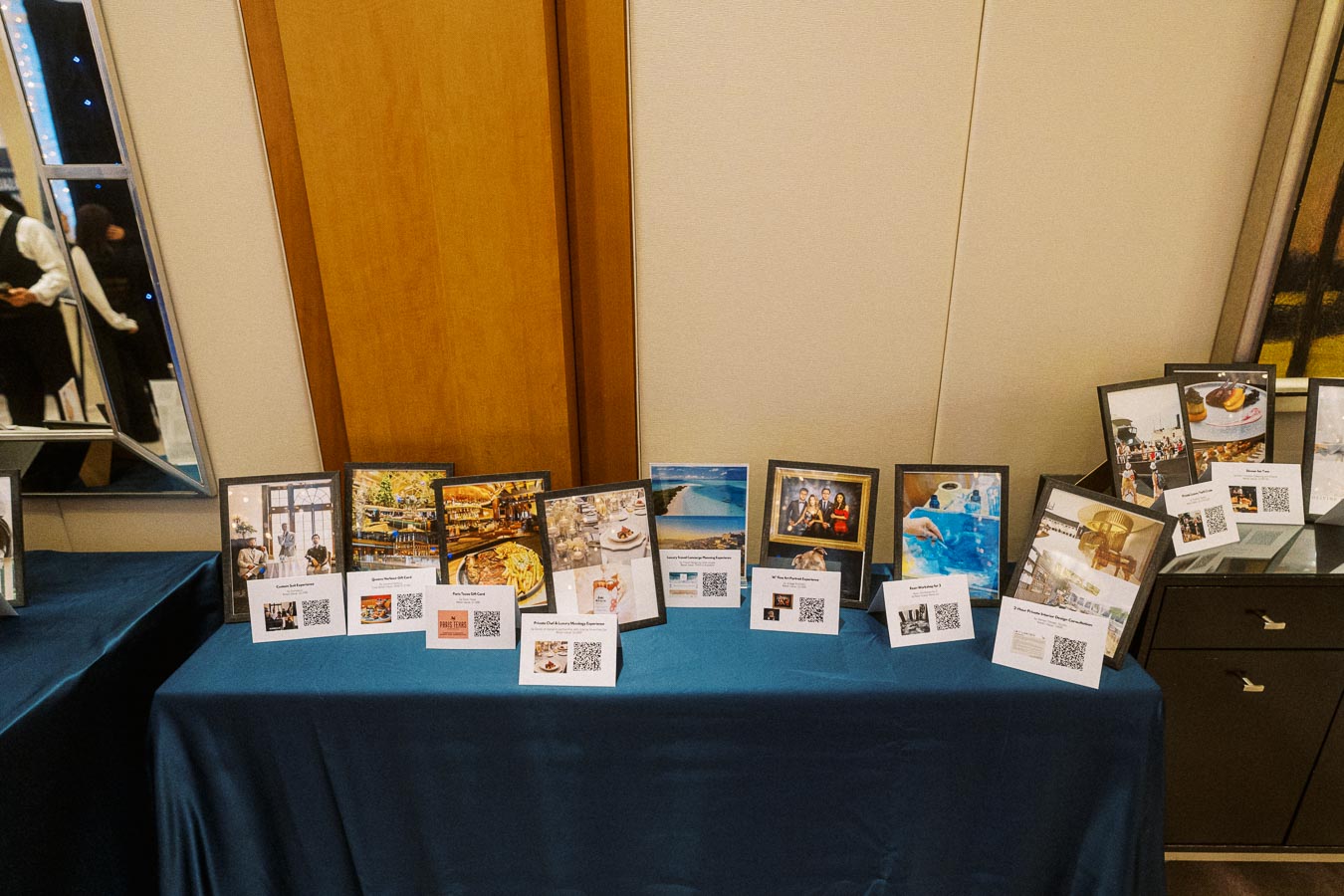 A display table at an event showcasing framed photographs and QR codes, indicating various travel destinations and experiences. The setting suggests an informative exhibition, possibly related to travel planning or destination marketing, against a backdrop of elegant decor.