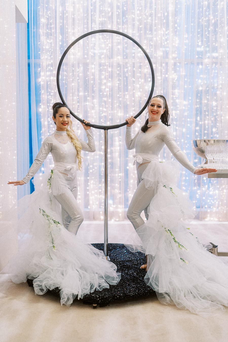 Two performers in elegant, shimmering costumes pose beside a large circular hoop against a backdrop of sparkling lights, creating a festive and glamorous atmosphere.