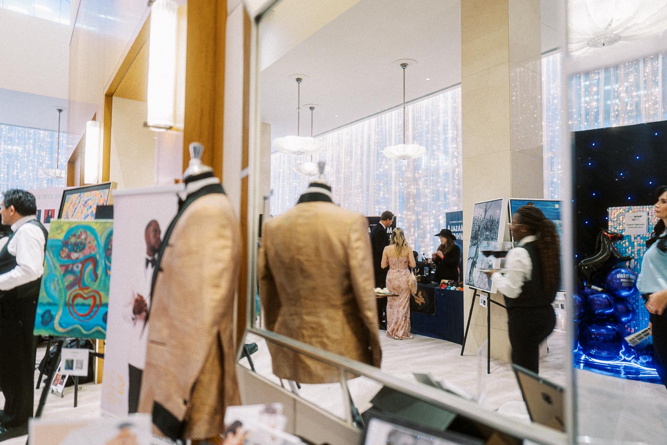 Elegant event showcasing art and fashion in a modern venue. Mannequins in stylish suits and colorful paintings are prominently displayed. Guests mingle in the background with decorative lighting creating a sophisticated ambiance.