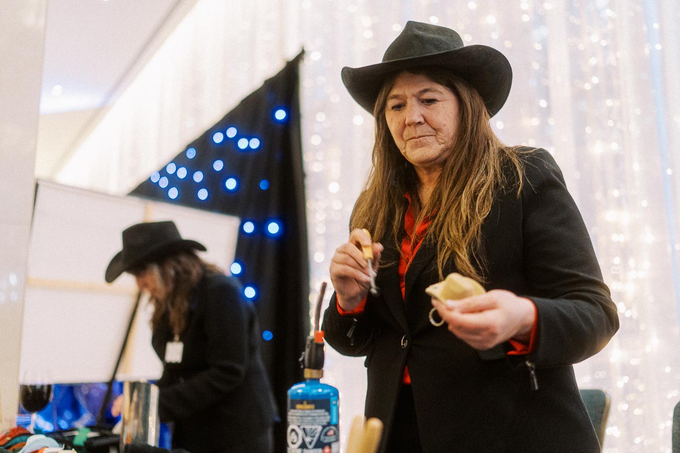A woman wearing a cowboy hat engages in a craft activity at an indoor event, with decorative lights in the background.