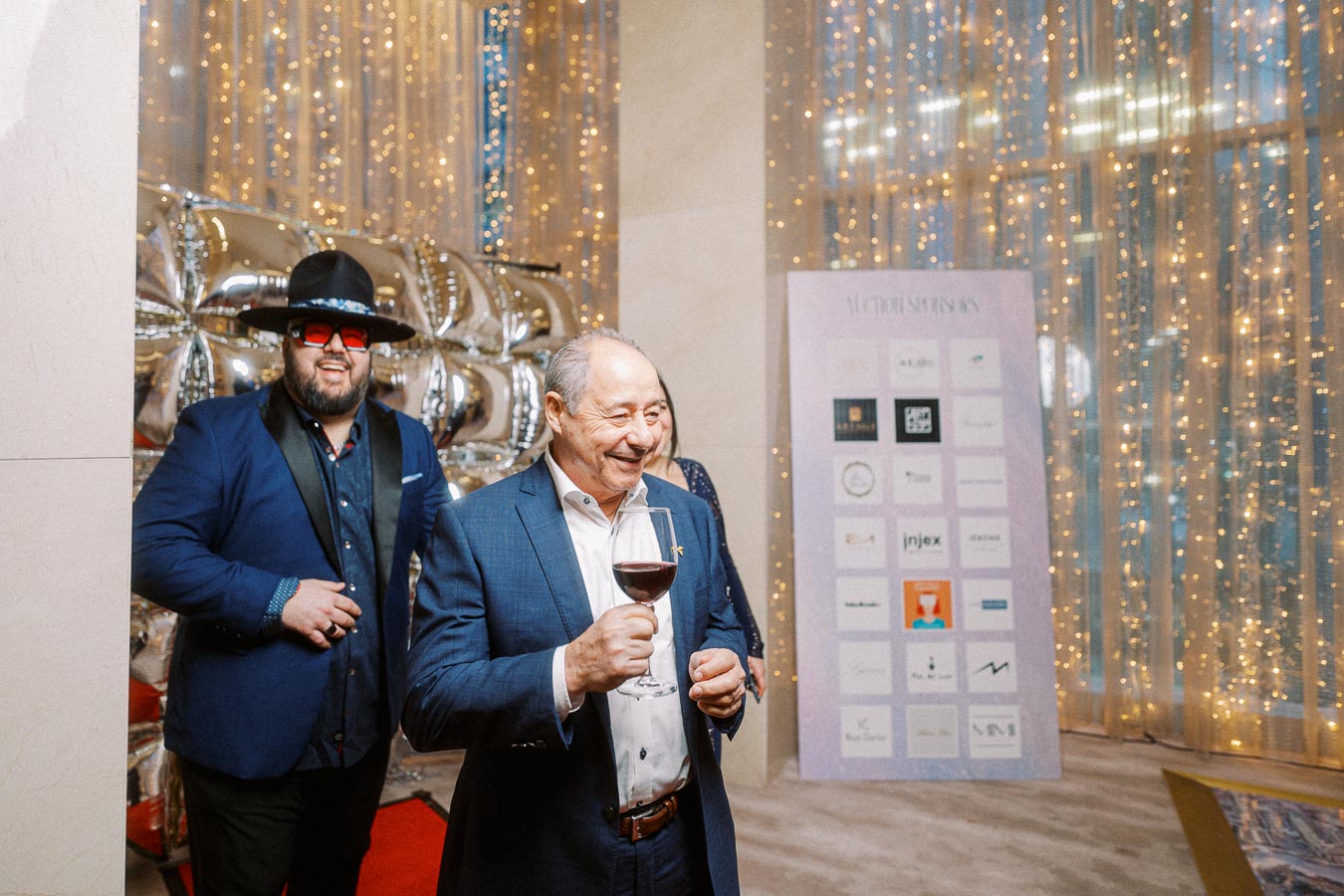Man in a blue suit holding a wine glass at a festive event, standing near a decorated wall with silver balloons and a board displaying logos, surrounded by warm lights.