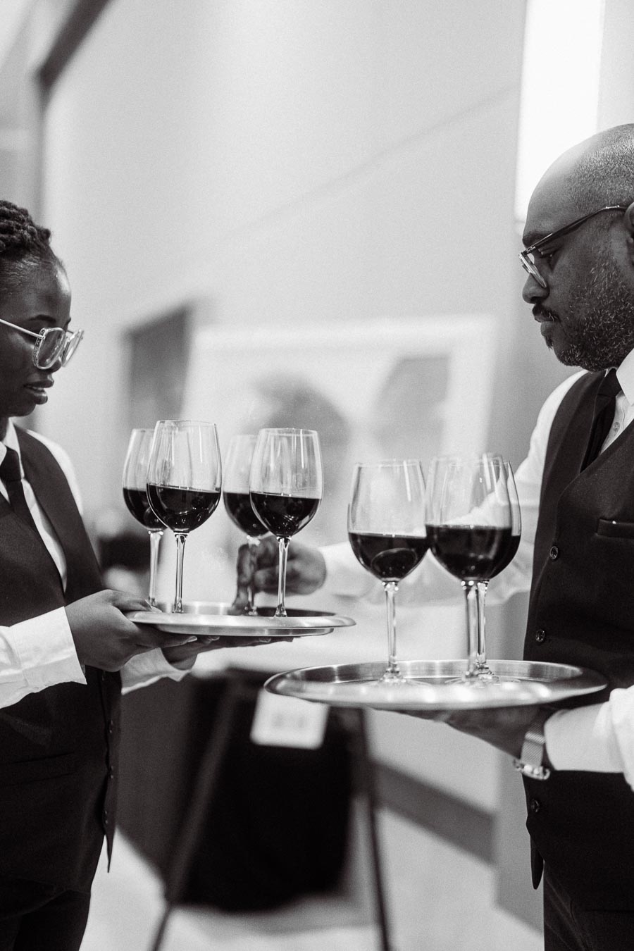 Black and white image of two servers holding trays with glasses of red wine at an event.