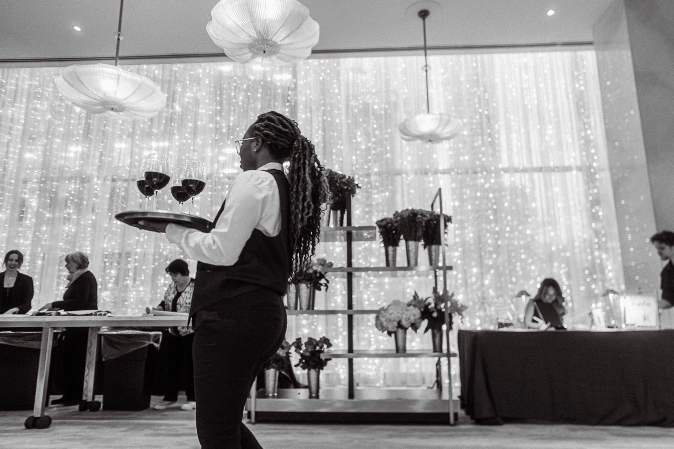 Elegant black and white photo of a waiter carrying wine glasses on a tray at a sophisticated event, with people and floral decor against a backdrop of twinkling lights.