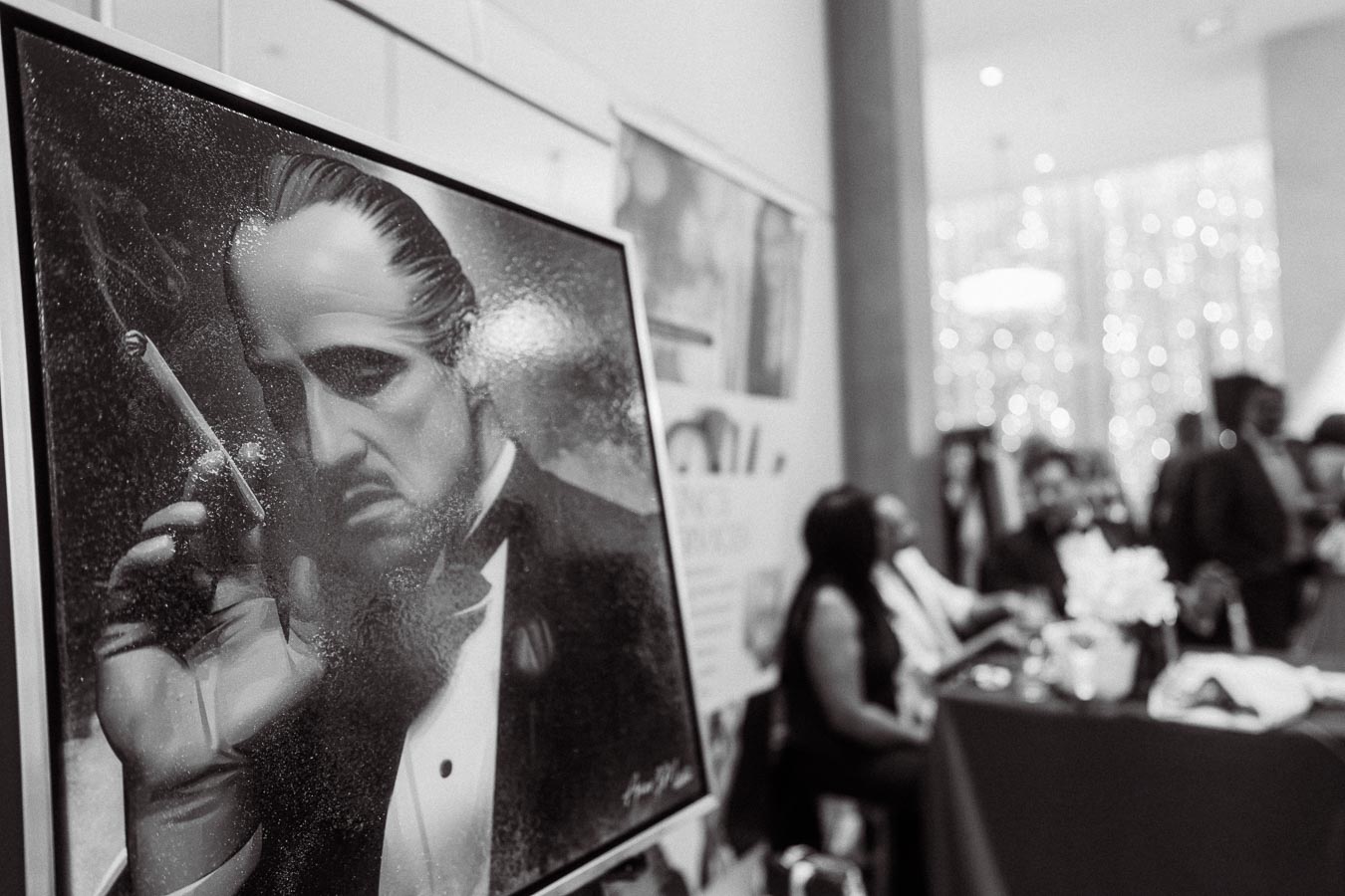 Black and white close-up of a painting depicting a serious man with slicked-back hair holding a cigar, featured prominently at a busy event with attendees conversing in the background.