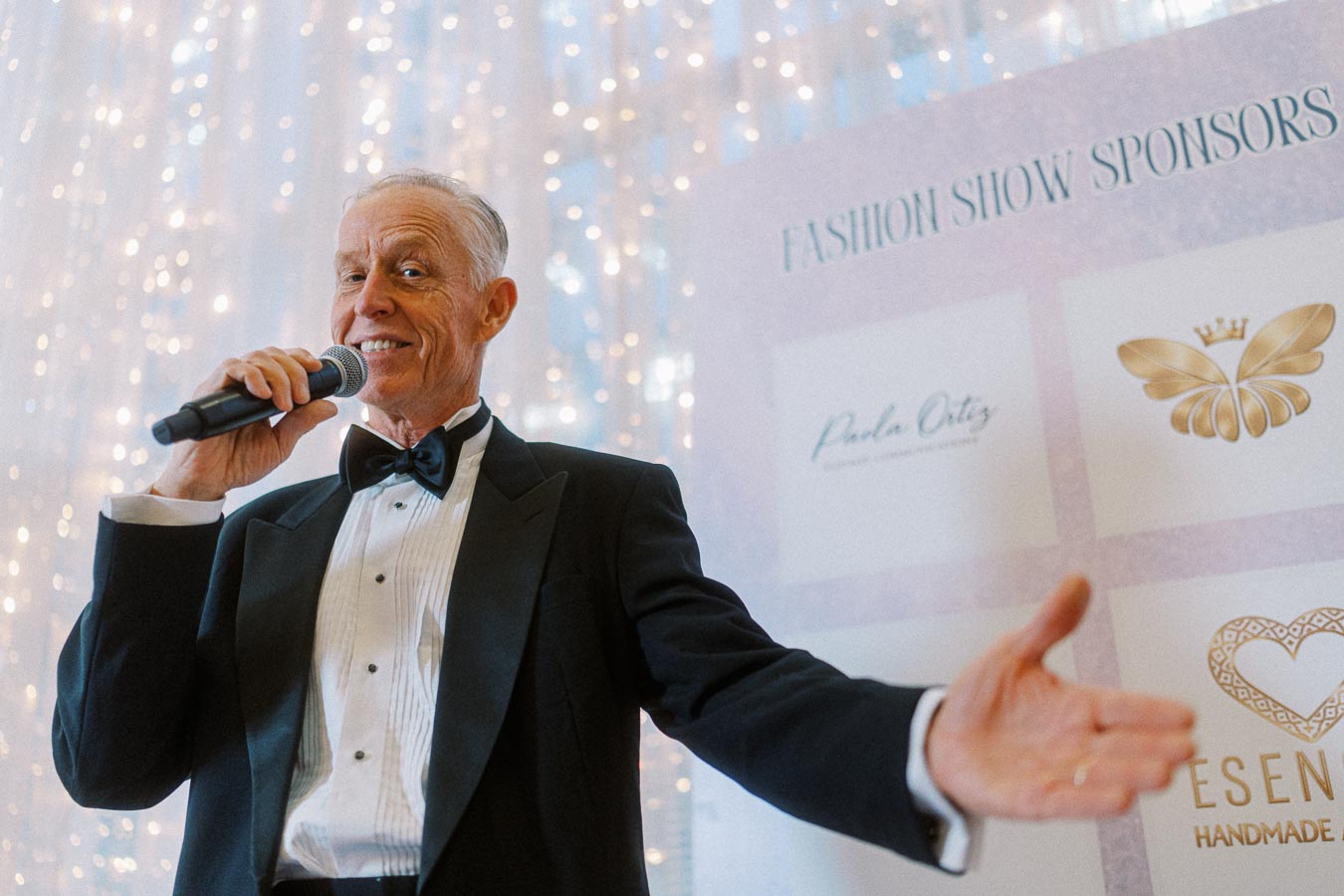 Elderly man in a tuxedo holding a microphone and speaking at a fashion show event, with a background of sparkling lights and sponsor logos.