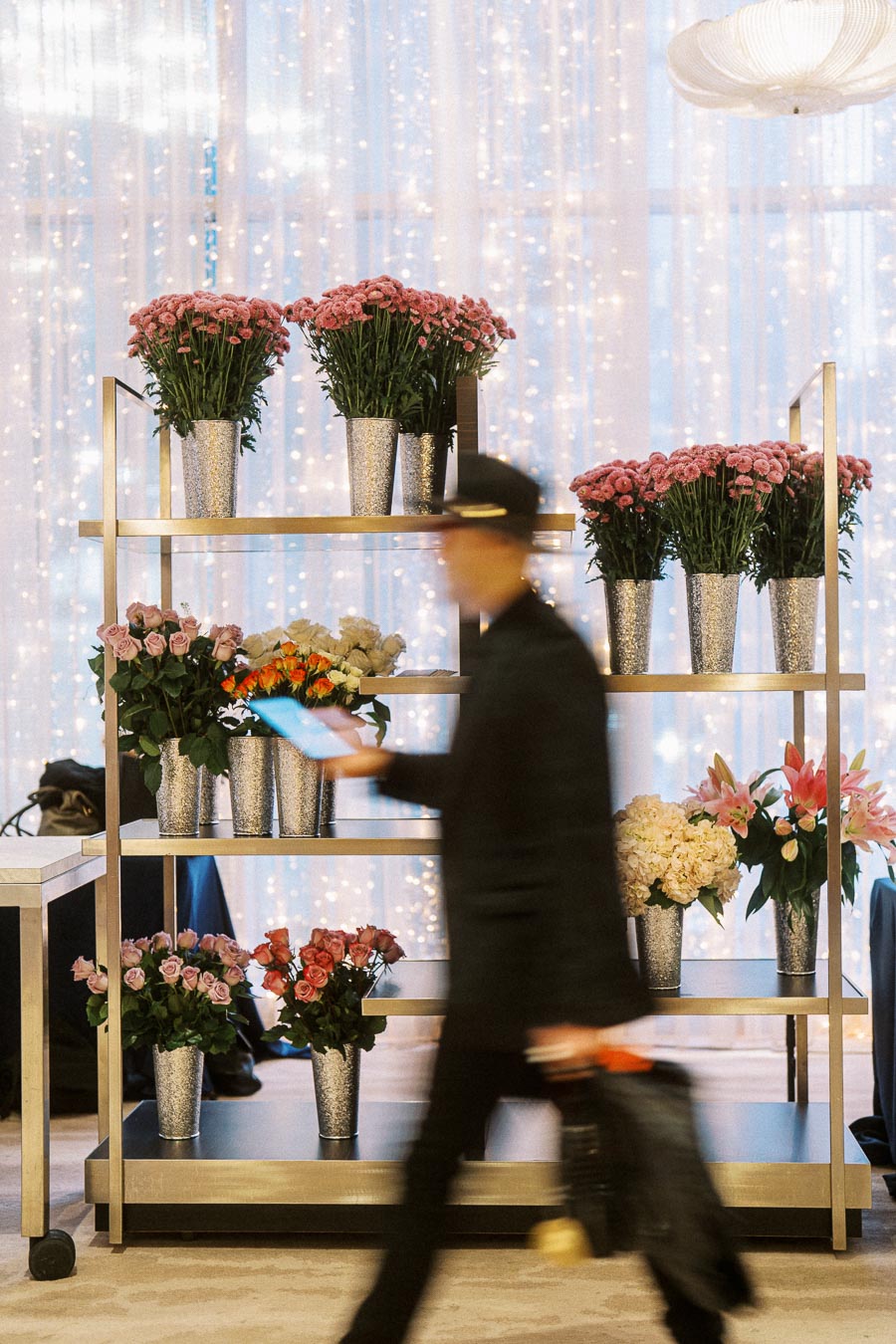 Flower display on metal shelves with numerous bouquets in vases, featuring pink carnations, roses, and lilies, set against a backdrop of twinkling lights. A person in motion, blurred, walks past the arrangement, adding a dynamic element to the elegant indoor floral setting.