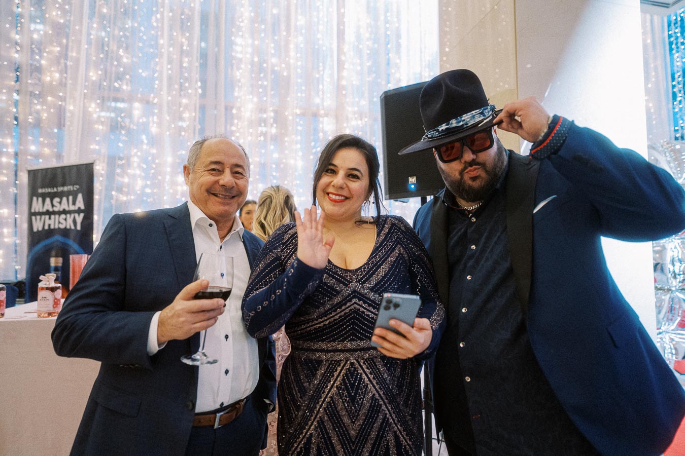 Three people enjoying a festive event, with sparkling lights in the background. A man in a suit holds a wine glass, a woman in an elegant dress waves with a smile, and another man poses stylishly with a hat. A Masala Whisky banner is visible, indicating a celebration atmosphere.