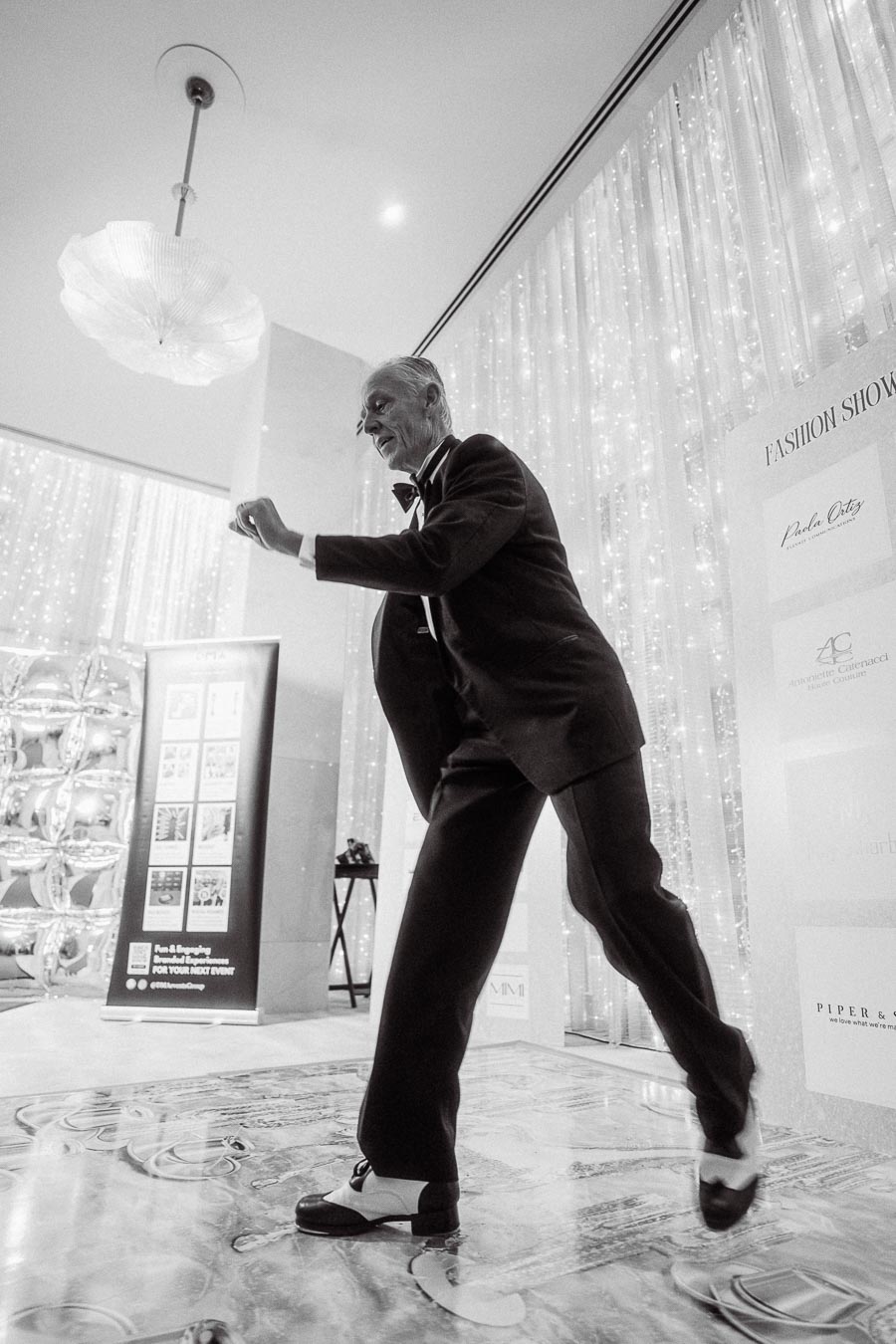 Elderly man elegantly tap dancing at a fashion show in a suit and bow tie, set against a backdrop of sparkling lights and event banners.