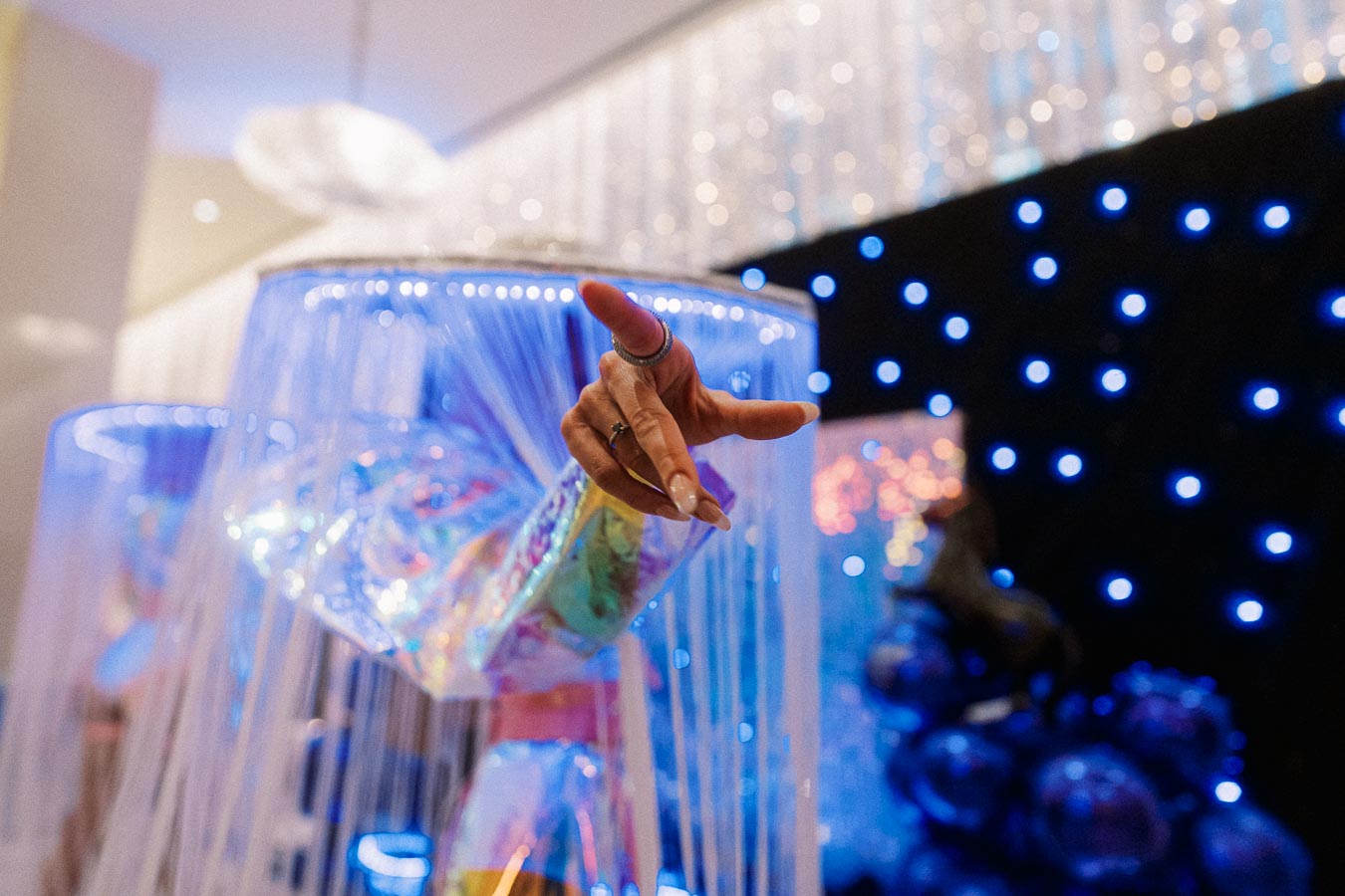 Iridescent futuristic costume with an outstretched hand, set against a backdrop of blue glowing lights and shimmering decor.