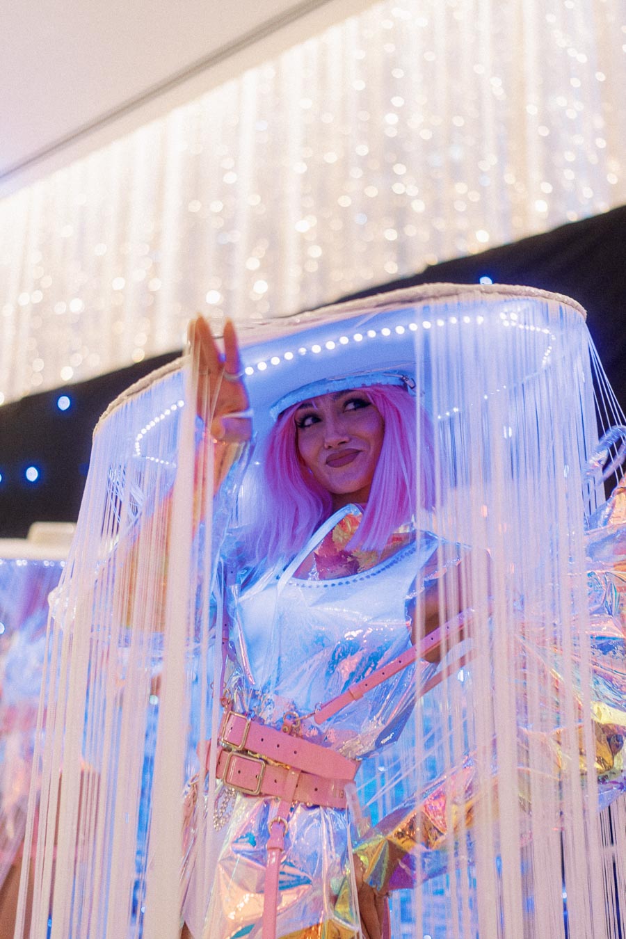 A person in a vibrant, futuristic costume with a pink wig and LED-lit accessories at a lively party.
