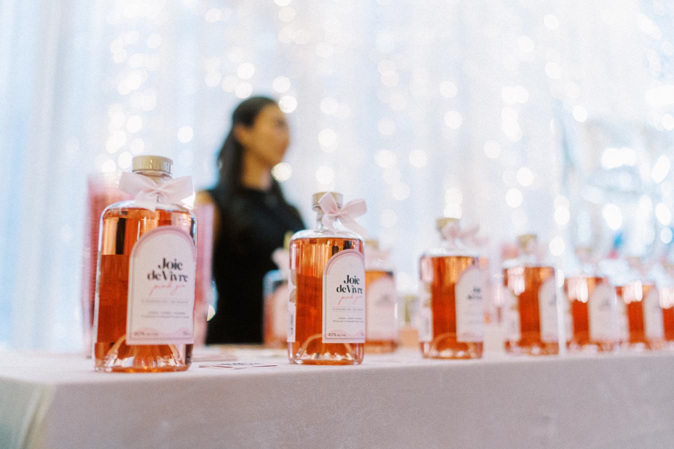 Bottles of Joie de Vivre pink gin displayed on a table with a blurred background, featuring a person standing among a softly lit, elegant setting.