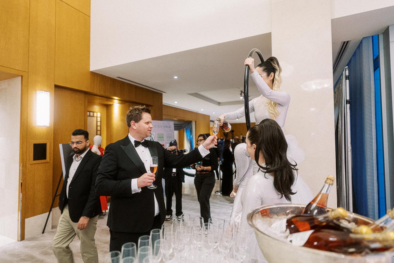 Elegant cocktail party scene with a man in a tuxedo receiving a drink from a performer on an aerial hoop, surrounded by guests and champagne bottles.