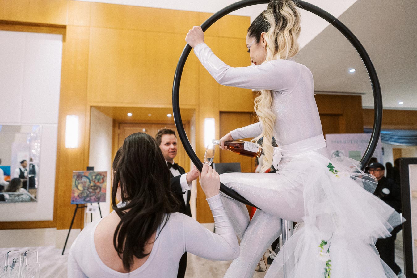 Aerial performer in a white costume pours drinks from a hoop at a sophisticated event, capturing the attention of elegantly dressed guests in a modern venue with art displays.