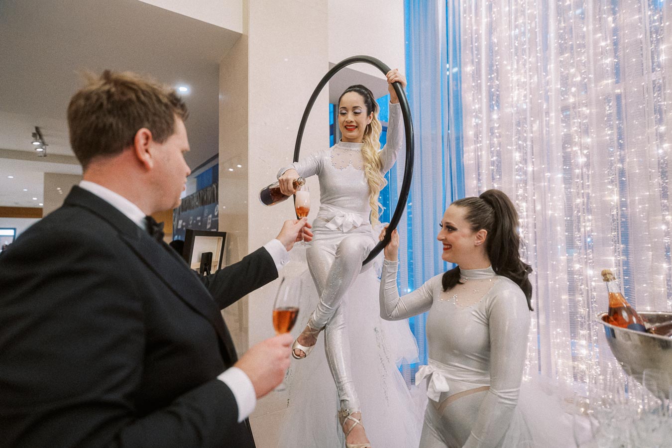 Aerial performer in a silver outfit pours champagne for a guest at an elegant event, accompanied by another performer, in front of sparkling blue and white curtain decor.