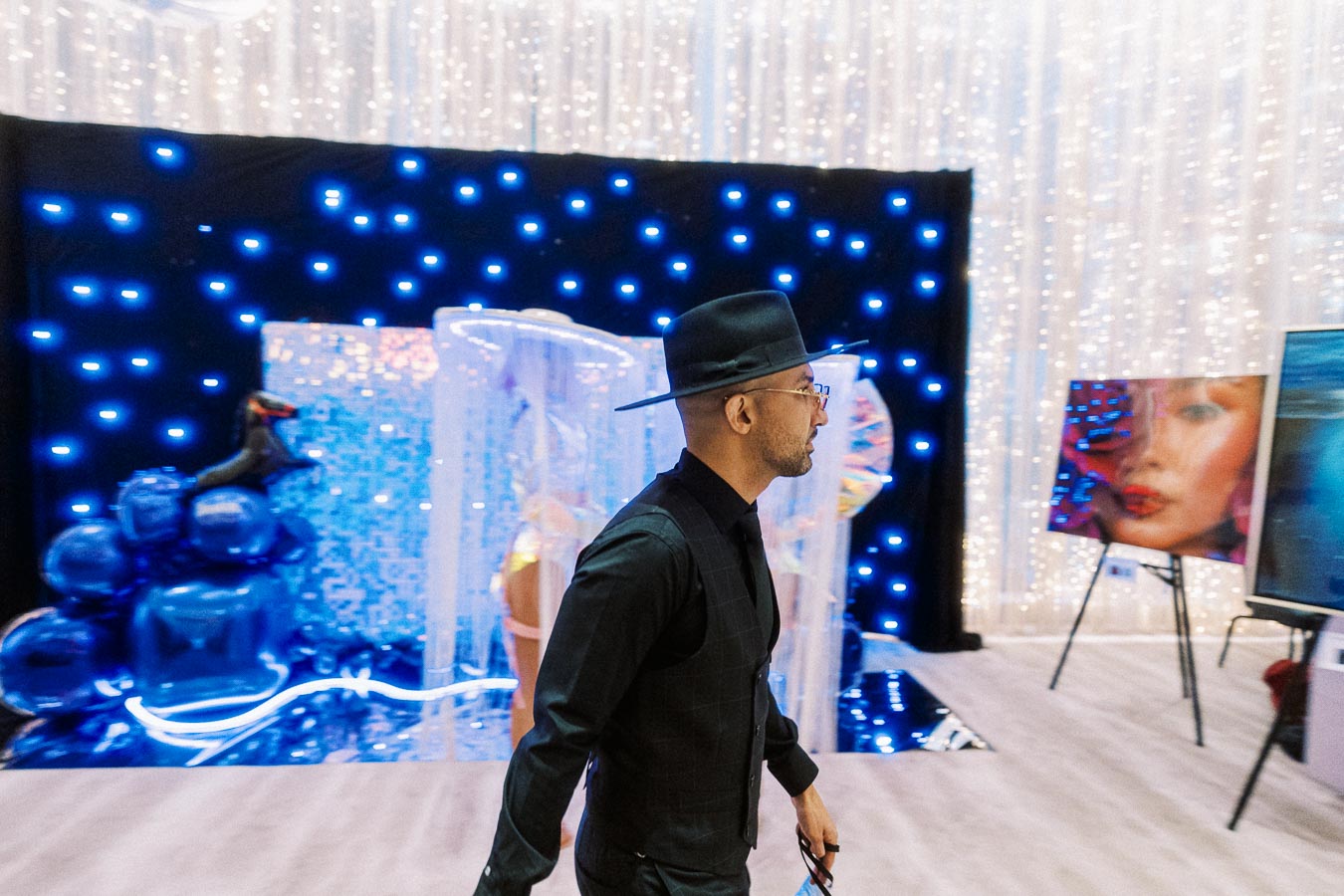 Man in a stylish black outfit and hat walking through a vibrant art exhibit with illuminated blue lights and colorful abstract paintings in the background.