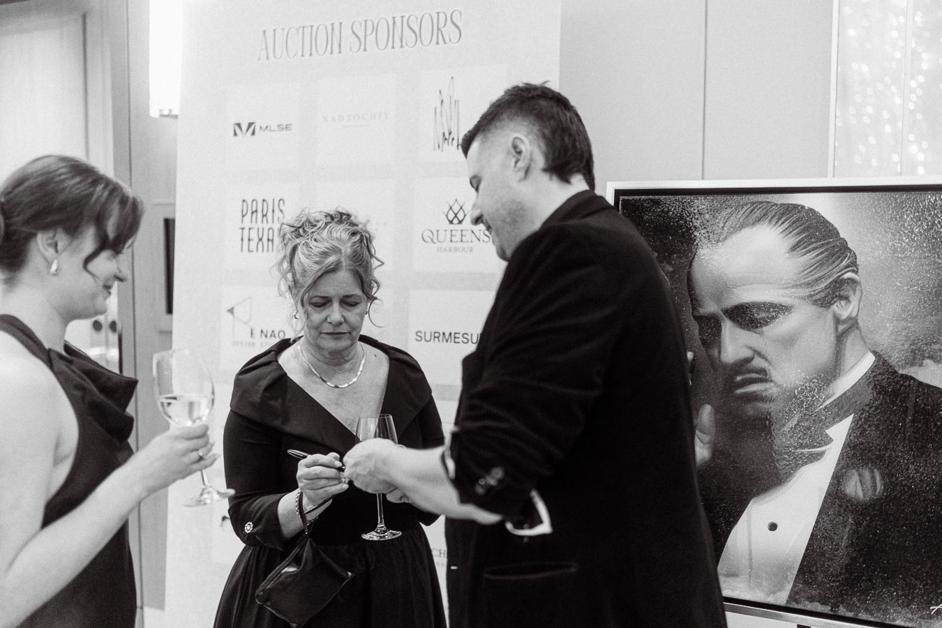 Black and white image of three people elegantly dressed at an auction event with wine glasses in hand. A large portrait resembling a famous film character is visible in the background. Signs with auction sponsors are displayed behind them.