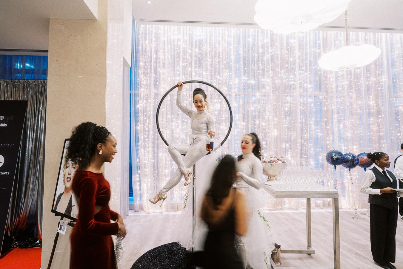 Aerial performer in a hoop entertaining guests at an elegant event with sparkling lights and balloons in the background.