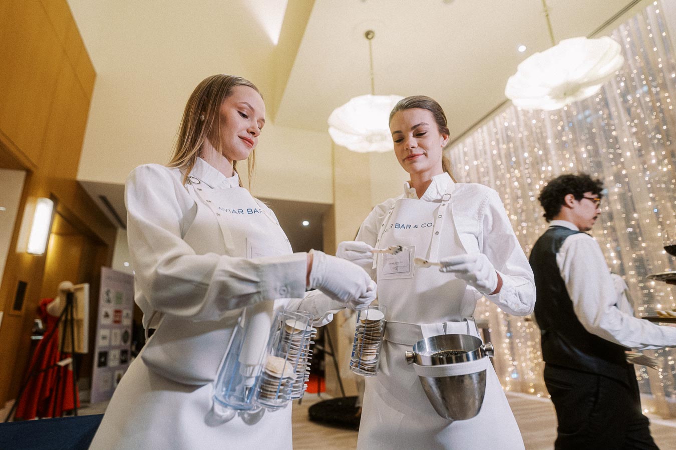 Two elegantly dressed women in white uniforms, preparing gourmet ice cream sundaes at a sophisticated event, with a beautiful backdrop of fairy lights in a modern venue.