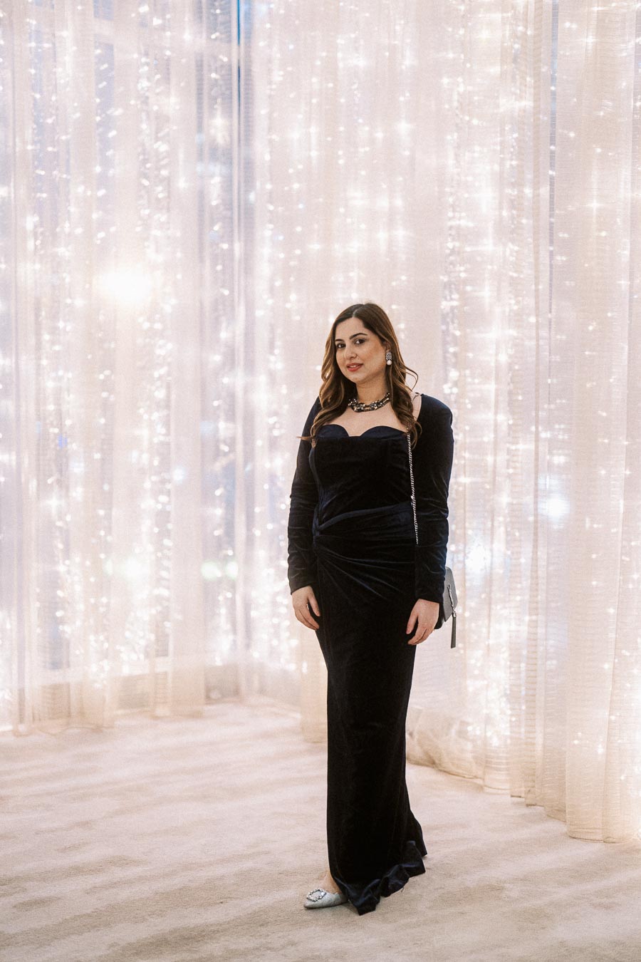 Elegant woman in a dark velvet gown standing in front of sparkling curtain lights, showcasing evening fashion and sophisticated jewelry.