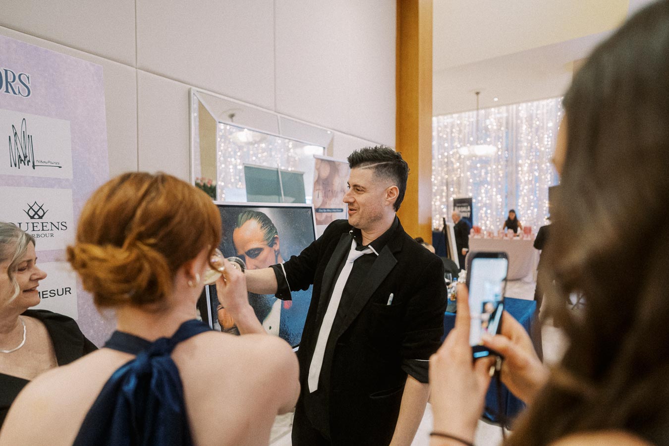 Man in a suit engaging with women at a formal event, one woman taking a photo with a smartphone, with elegant decor and branded signage in the background.