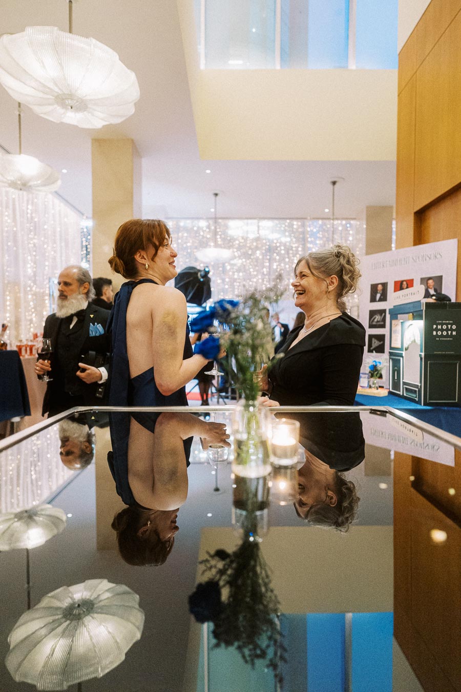 A lively gala scene with two women engaged in conversation near a reflective surface, surrounded by elegant decor and soft lighting, capturing the festive and sophisticated atmosphere.