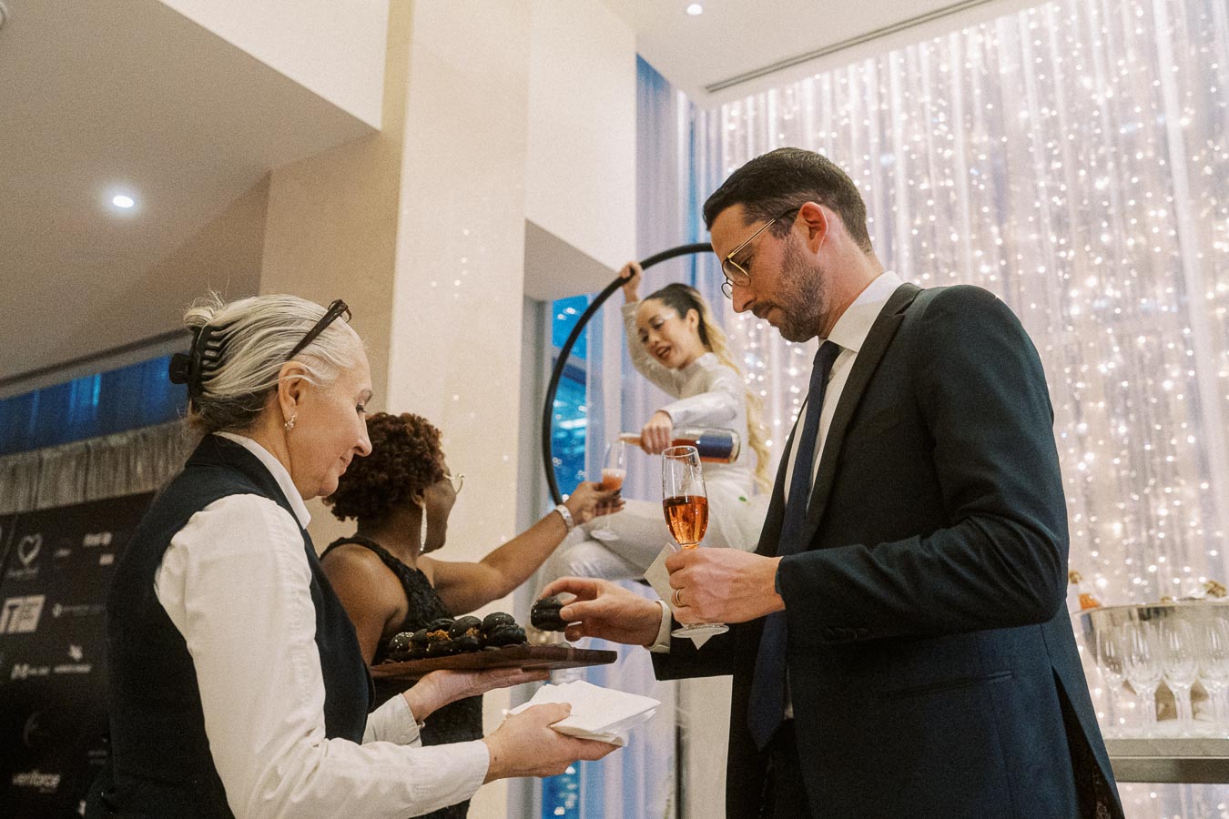 Elegant corporate event with attendees enjoying hors d'oeuvres and champagne, featuring a performer pouring drinks from a hoop, against a backdrop of shimmering lights.