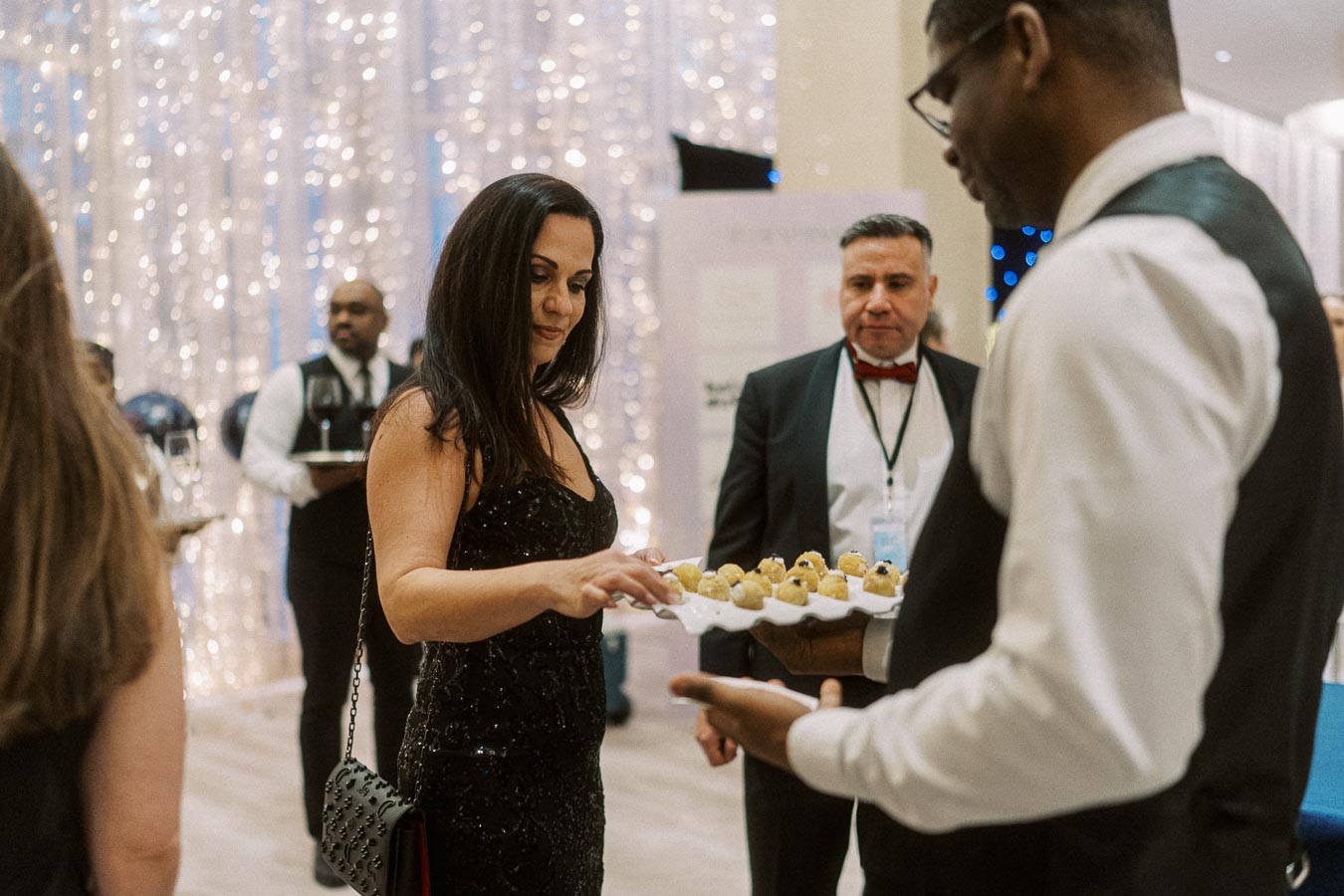 Elegant gala event with a woman in a black dress selecting hors d'oeuvres from a server's tray, surrounded by guests in formal attire against a sparkling backdrop.