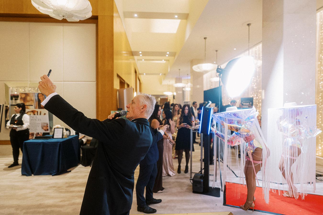 Elderly man taking a selfie at a glamorous event with elegantly dressed attendees and performers in vibrant costumes on a red carpet in a bright, luxurious venue.