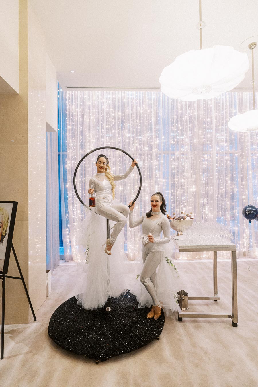 Aerial performer sitting gracefully on a circular hoop, smiling and holding a drink, next to an elegantly dressed woman, standing in front of sparkling curtain lights at a sophisticated event setup.