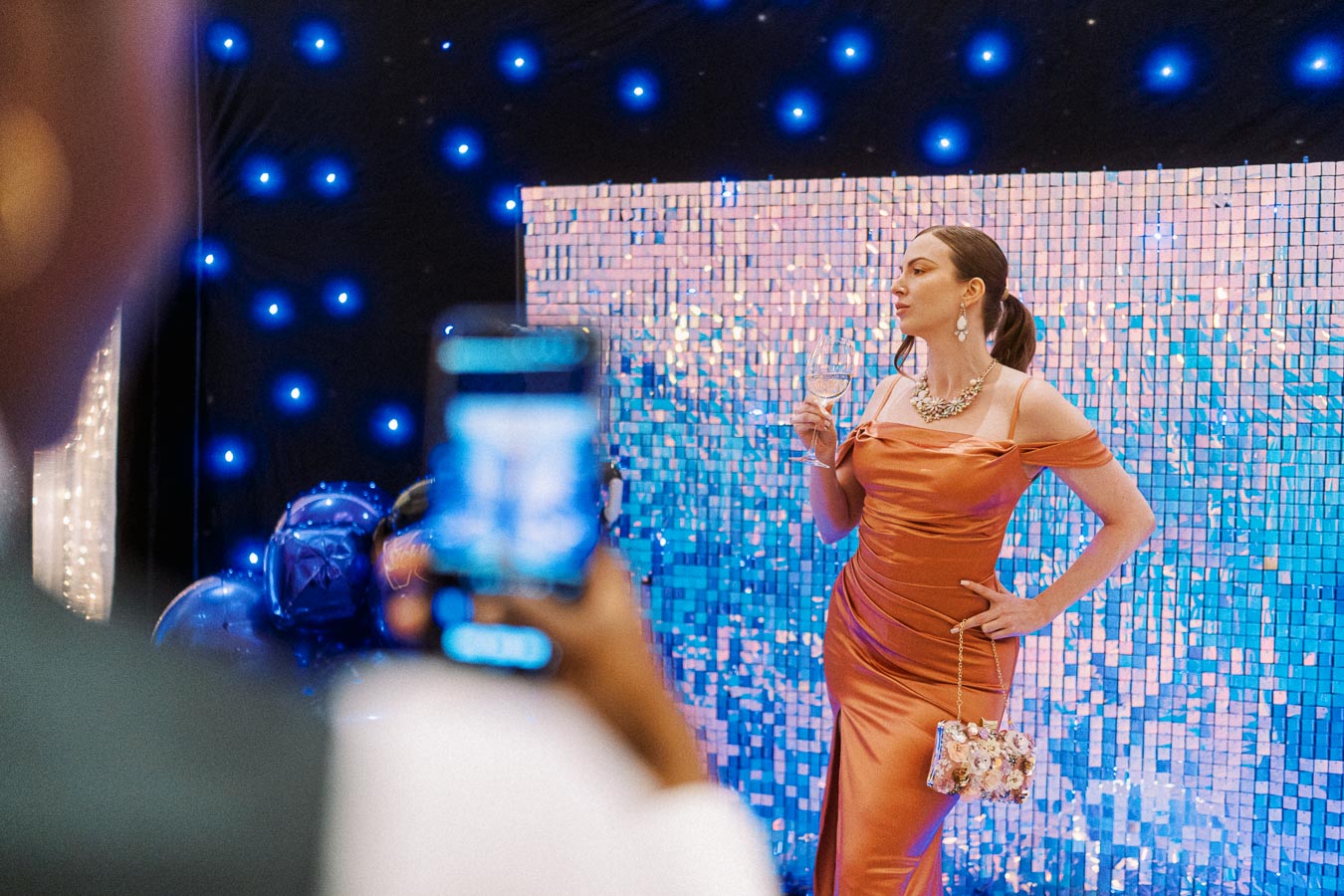 Woman in an elegant orange dress holding a wine glass, posing at a glamorous event with a sparkling sequin backdrop and blue lights captured on a smartphone camera.