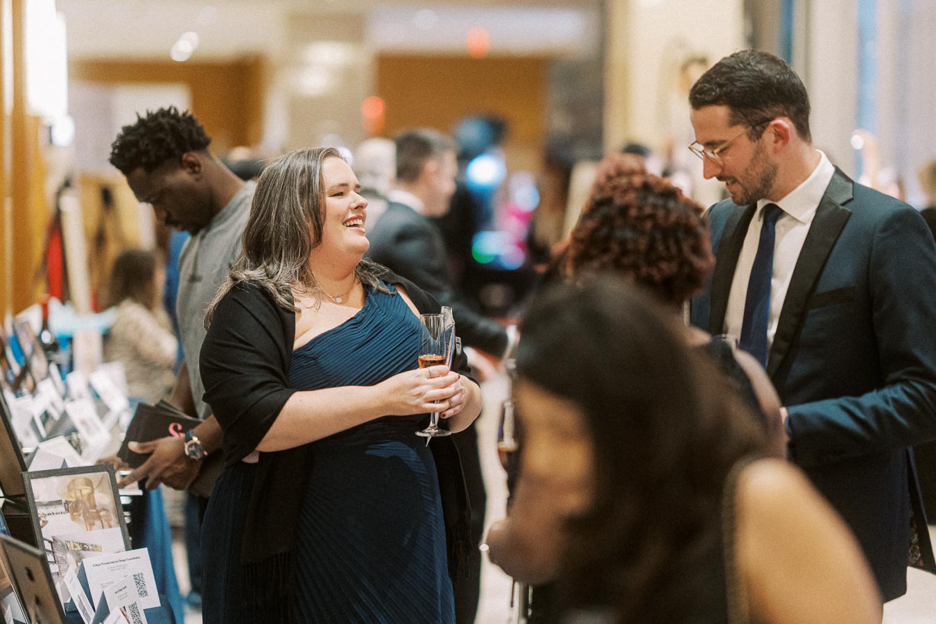 A joyful group of people in formal attire interacting and networking at an elegant event, with tables showcasing various items in a warmly lit room atmosphere.