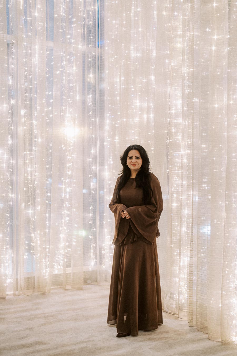Elegant woman in a long brown dress standing in front of sheer curtains adorned with twinkling fairy lights, creating a sophisticated and magical atmosphere.