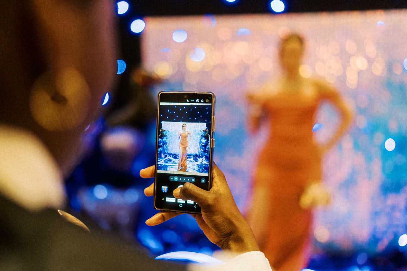 Person capturing a fashion model on stage with a smartphone, against a colorful, blurred backdrop. The model is in focus on the phone screen, showcasing the vibrant atmosphere of the event.