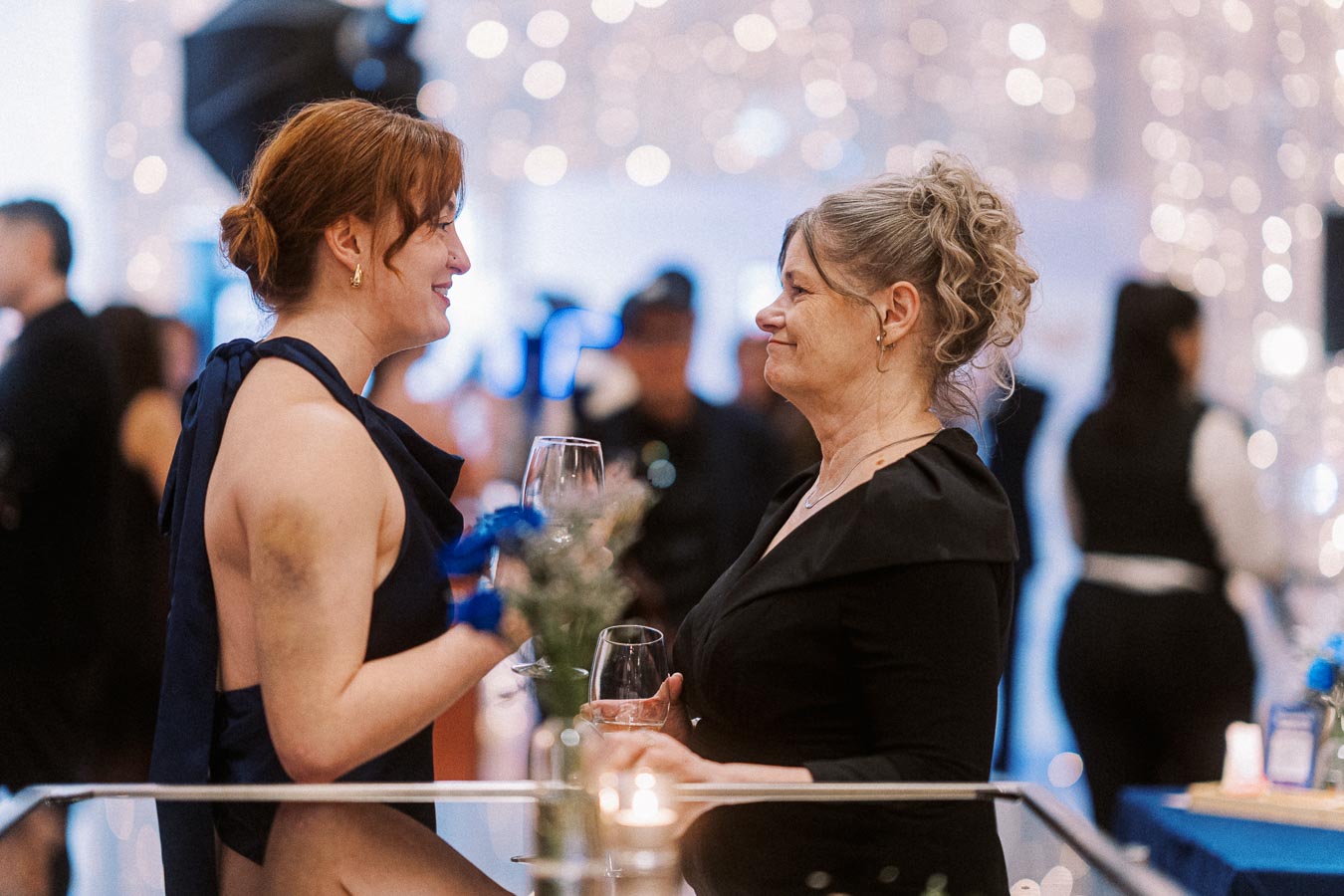 Two women in elegant attire conversing and smiling while holding champagne glasses at a festive gathering, with a blurred background of lights and guests.
