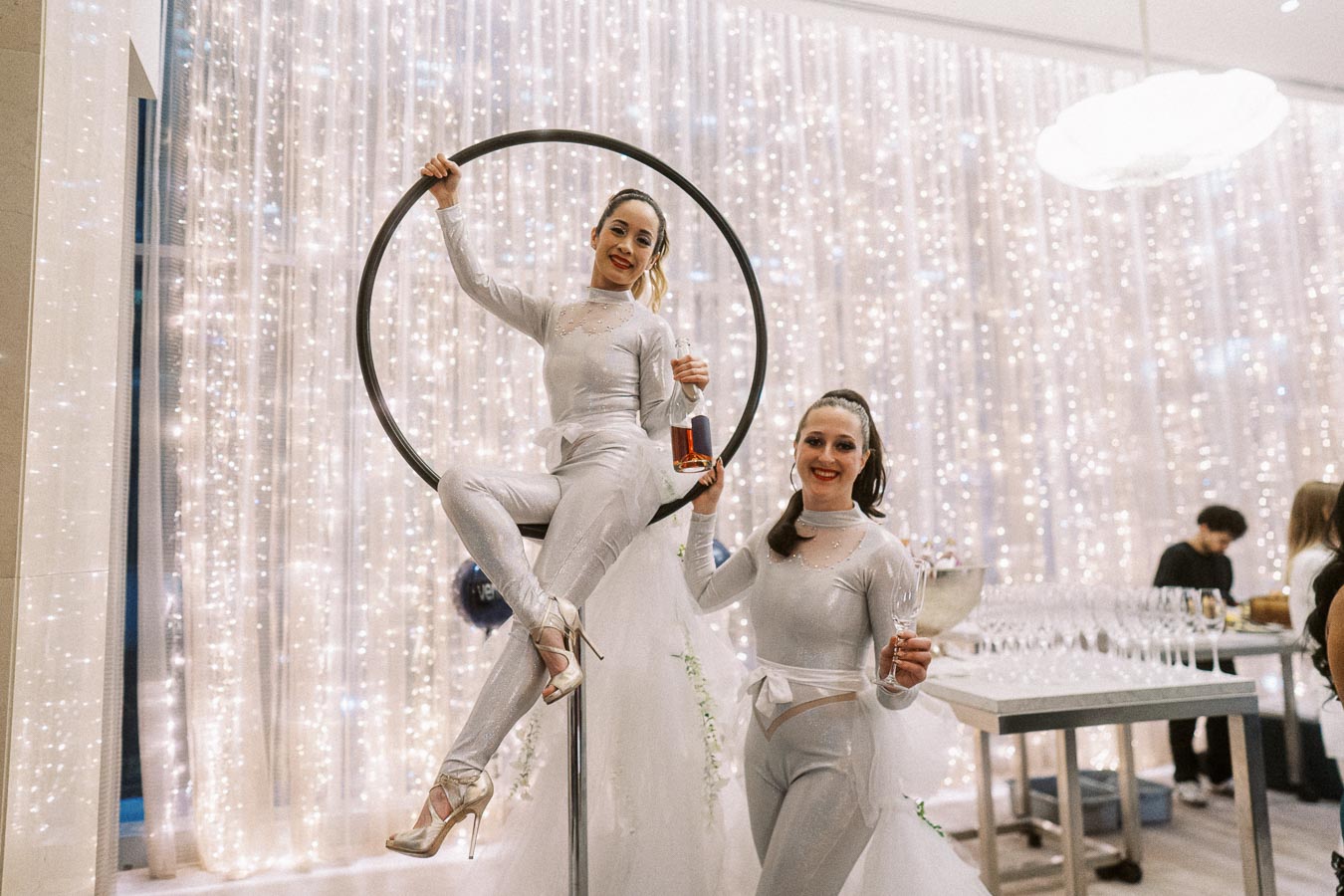 Two performers in elegant silver costumes pose at a glamorous event, with one seated on an aerial hoop holding a bottle, and the other standing with a champagne glass, in front of a backdrop of twinkling lights.
