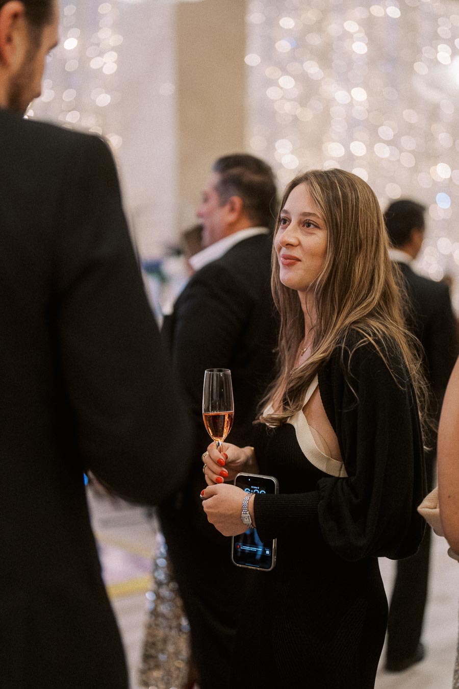 Elegant woman holding a glass of champagne at a sophisticated event, surrounded by people in formal attire, against a backdrop of sparkling lights.