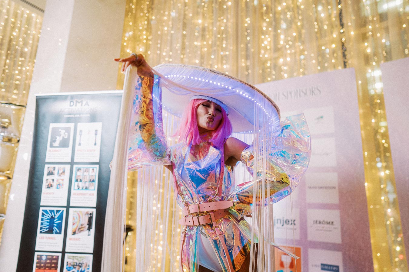 Person in a vibrant, iridescent costume with pink hair and a large, futuristic hat, striking a pose in front of a backdrop with golden lights and event signage.