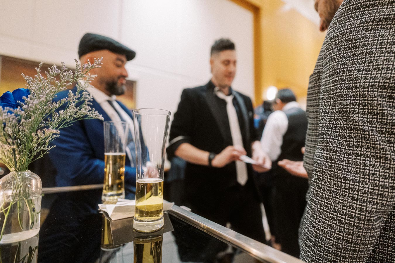 Elegant social gathering with people in formal attire, featuring a focus on a glass of champagne and decorative flowers.