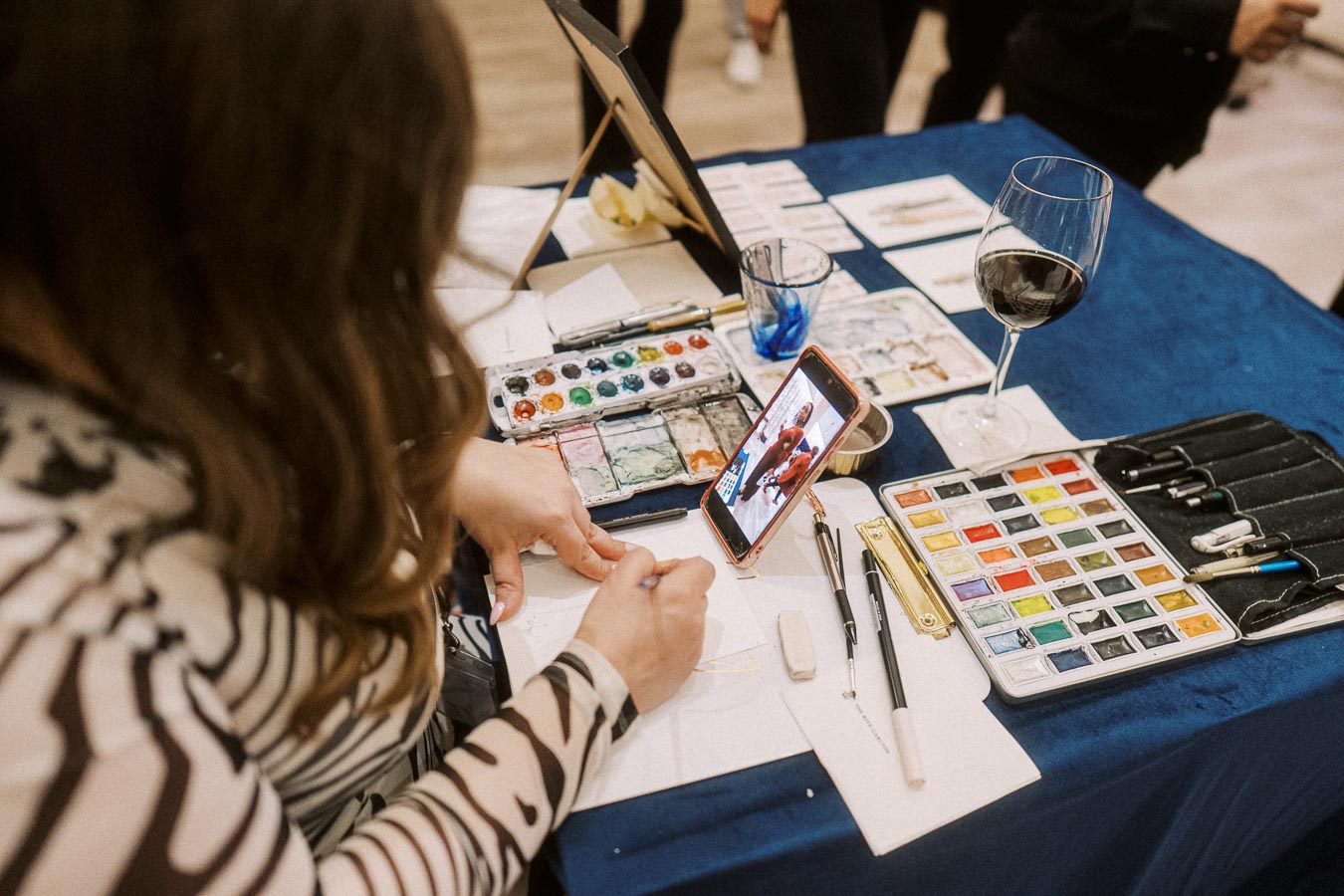 Artist creating a watercolor painting with reference on smartphone, surrounded by art supplies and a glass of wine on a blue cloth-covered table.