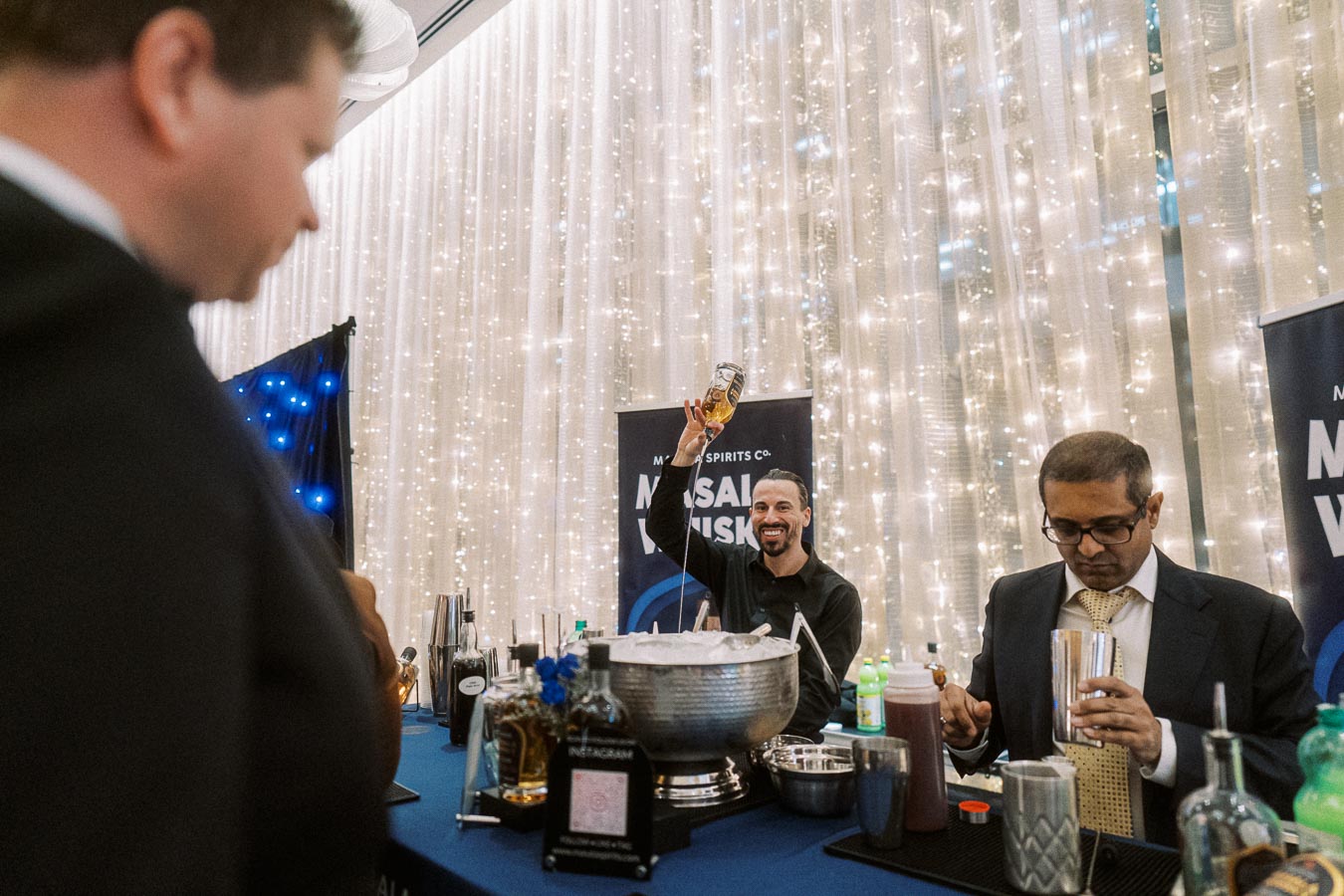 Bartender preparing drinks at a lively event with decorative lights in the background, featuring a booth promoting Mosaic Whisky.