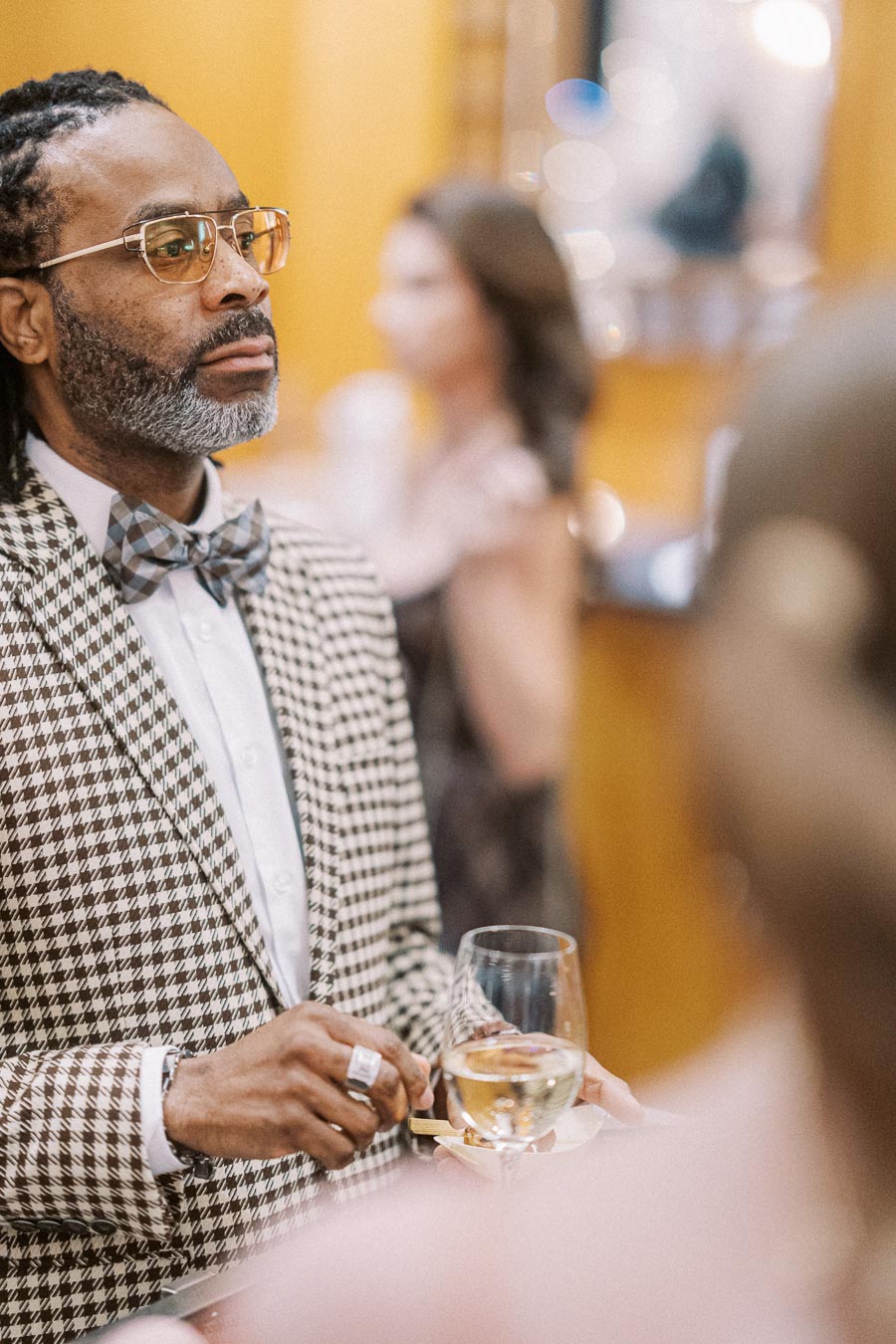 Stylish man in a checkered suit and bow tie, holding a wine glass at a formal event.