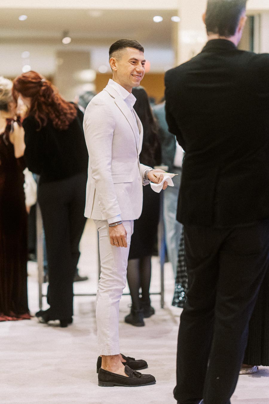 Man in a light gray suit smiling at a formal event with people socializing in the background.