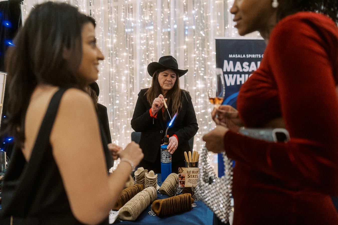 Guests enjoying a whiskey tasting event with a variety of branded spirits on display, while a woman in a hat uses a torch for a demonstration, surrounded by festive lights.