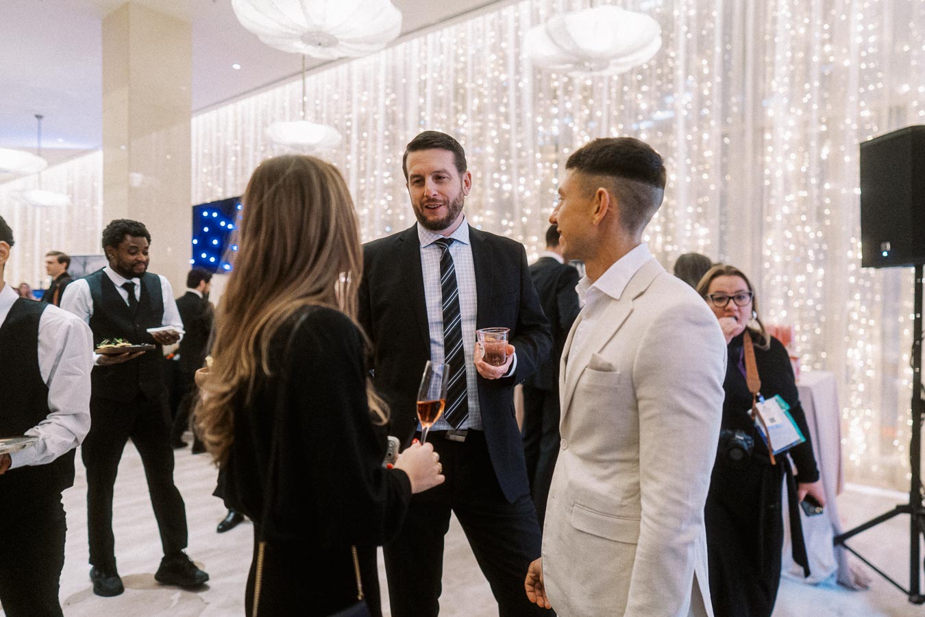 A group of well-dressed people engaged in conversation at an elegant event with sparkling lights in the background. A waiter serves appetizers, and the atmosphere is lively and sophisticated, suggesting a formal gathering or celebration.