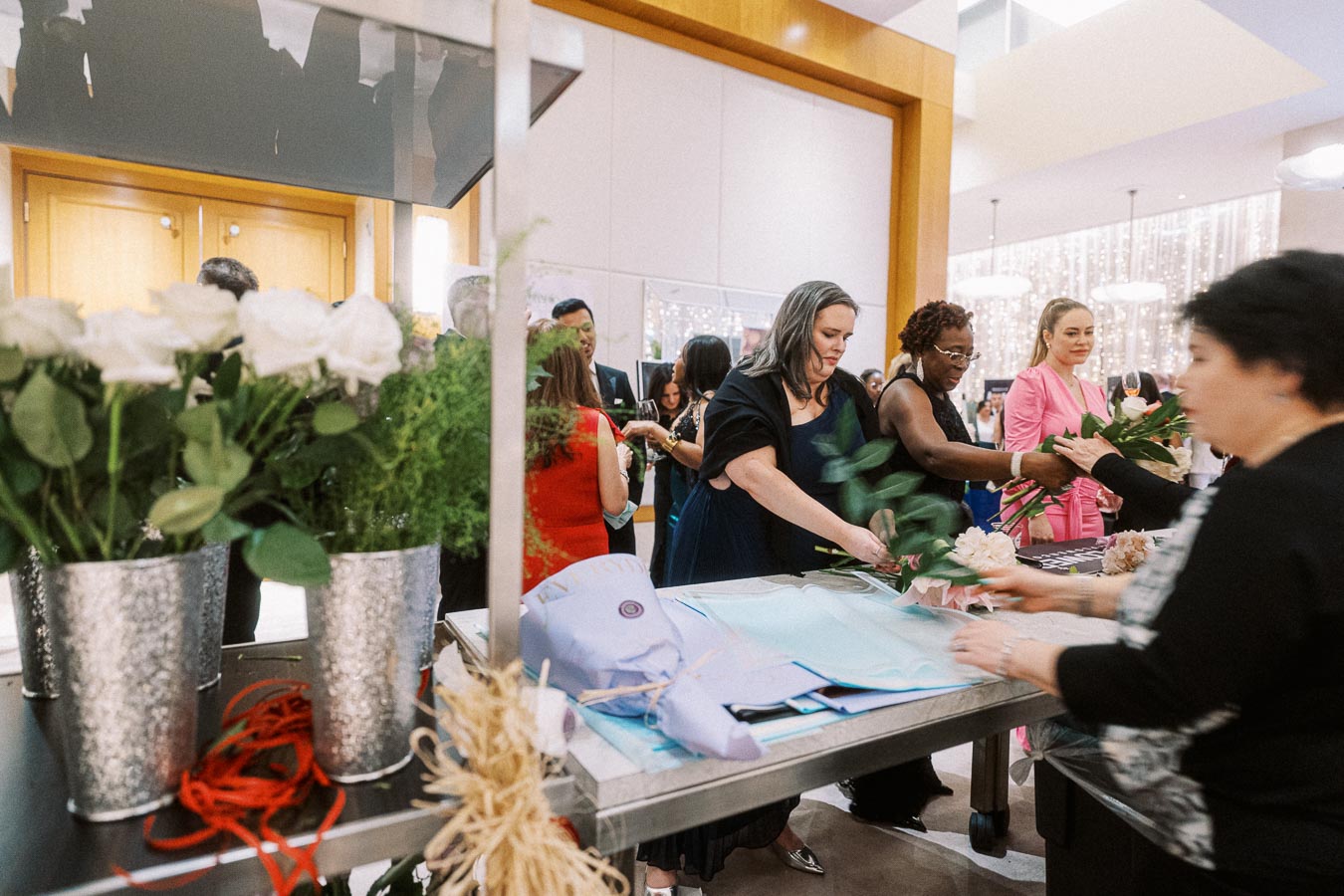 Group of people arranging flowers at an event, with white roses in metallic vases on a table.