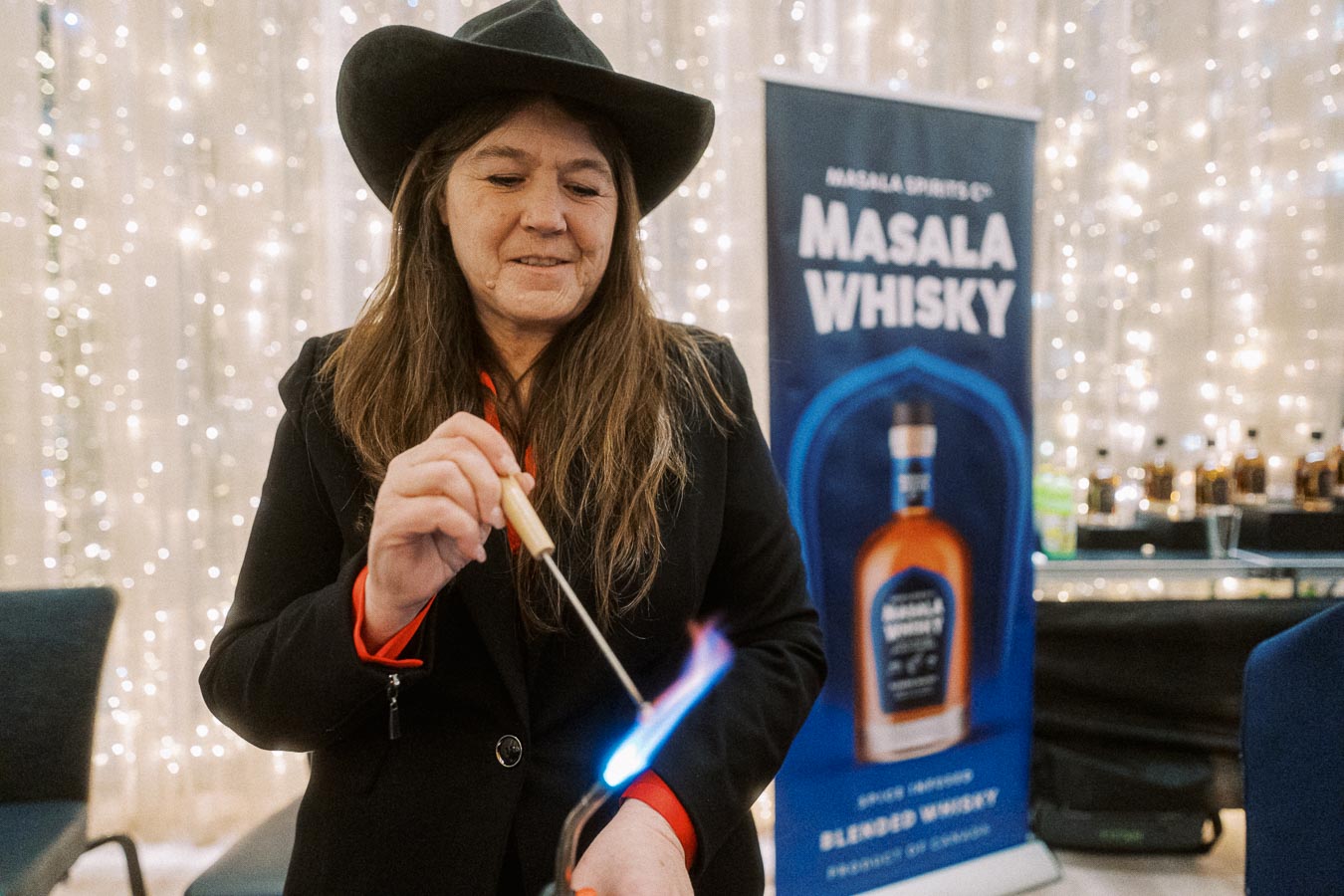 A woman in a cowboy hat ignites a small flame at a whisky tasting event with a Masala Whisky banner in the background.