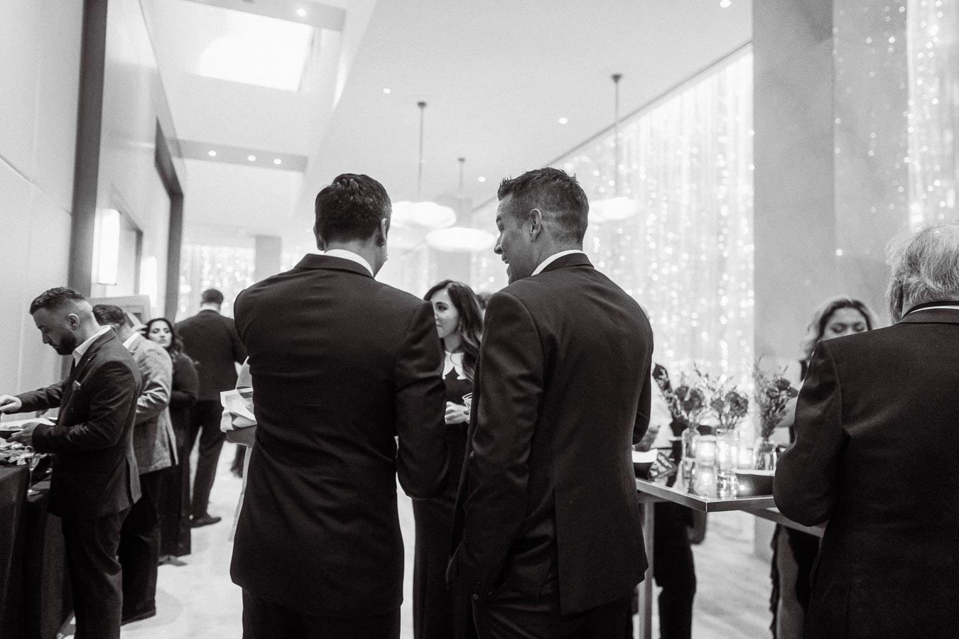 Black and white image of a group of people in formal attire engaging in conversation at a sophisticated networking event. The setting features elegant decor with soft lighting, creating a refined atmosphere. Ideal for business and corporate event themes.